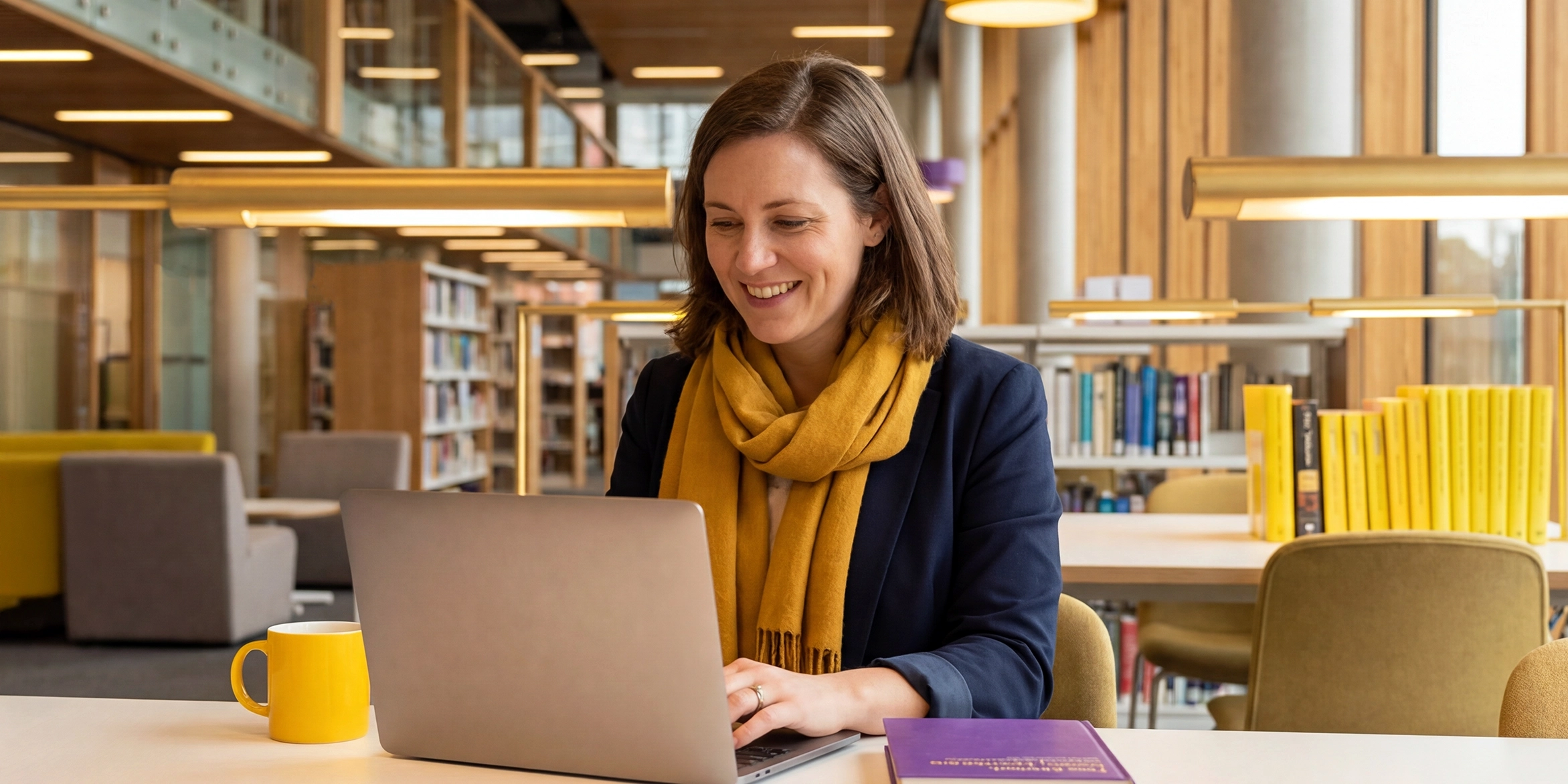 A library professional sitting at a desk in a academic library, working on a laptop.