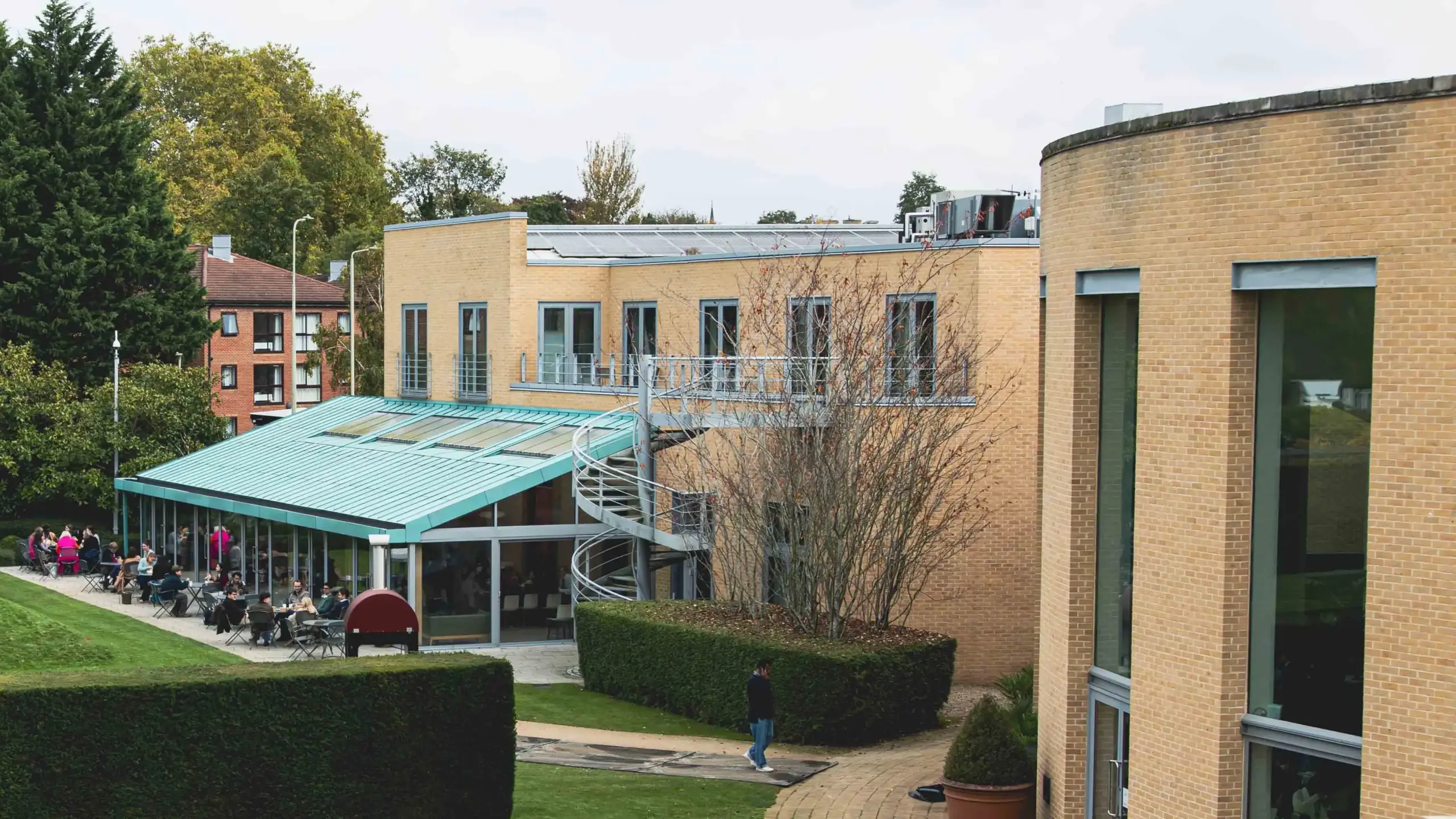 An aerial view of the Saïd Business School campus showing a courtyard café area with a teal canopy, surrounded by brick buildings and green lawns.